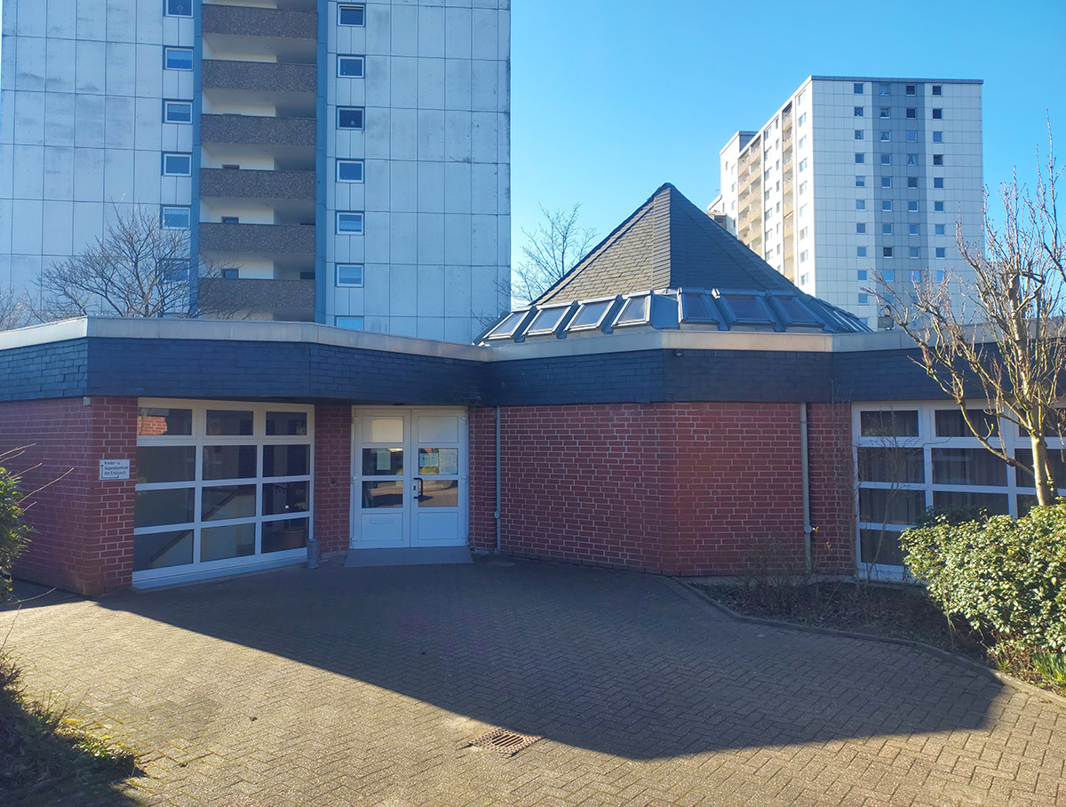 Moderner Backsteinbau mit Zeltdach vor zwei großen Wohnblocks.